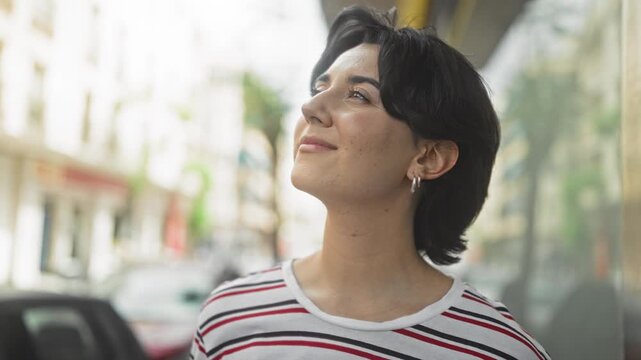 Young woman looks up, face and hoop earring visible on a street with glass storefronts, wearing a striped shirt; serenity reflection.