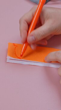 Adult hand draws a face with jagged teeth and a triangle eye using an orange felt tip pen on a folded napkin over a pink surface, a simple craft idea.