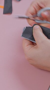 Female hands press a folded black paper napkin flat and snip small notches with slim manicure scissors on a pink surface, a step by step home craft lesson for spooky party decor.