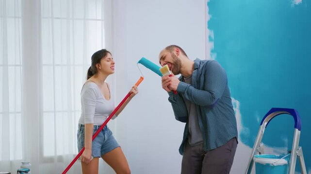 Man uses a paintbrush as a pretend microphone while a woman laughs with a long roller, showing playful teamwork during apartment redecorating.
