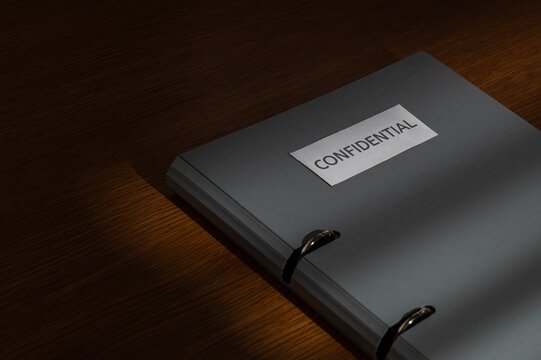 Gray ring binder labeled CONFIDENTIAL on a wooden desk. Corporate secrecy, classified documents, legal privacy concept.