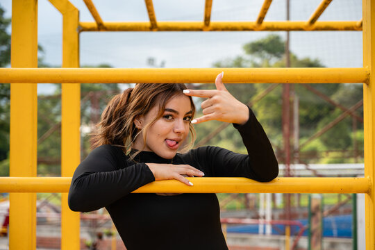 Colombian teenager showing playful expression with tongue out and rock and roll hand gesture, having fun on yellow monkey bars at a park playground during daytime
