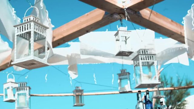 White decorative lanterns hanging from wooden beams against a clear blue sky with flowing white fabric draping overhead
