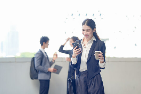 A smiling Asian businesswoman in a dark suit reading an email on a smartphone during a coffee break on a skywalk above the business district, showing how work happens beyond the meeting room