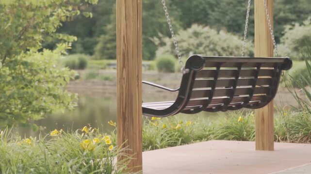 Black metal park swing hanging between wooden posts beside a pond with yellow flowers, calm