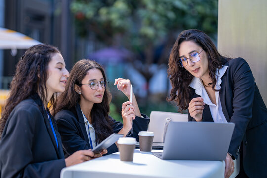 A diverse team of three Hispanic businesswomen in dark suits, deep in an animated discussion, hold a team meeting at an outdoor cafe table pointing at the laptop screen.
