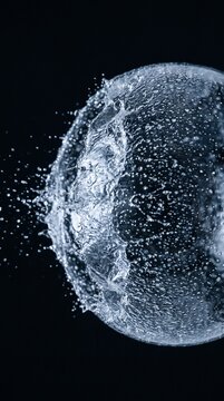 High-speed photograph capturing a spherical water balloon bursting into countless droplets against a dark background.