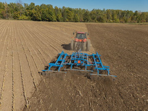 Gregorville, Wisconsin, United States - 05 October 2026: Aerial view of a red tractor tilling a large field with blue agricultural equipment under a clear sky.