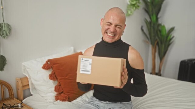 Bald man holding cardboard package on bed, smiling broadly while lifting a shipping box toward camera in a studio bedroom; joyful unboxing.