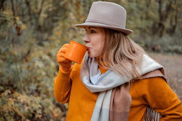 Fototapeta premium Woman drinking hot tea from mug in autumn park - cozy seasonal outdoor moment and warming beverage concept. Profile view of female in hat drinking coffee outdoors.