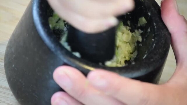 Hand grinding garlic and green onions in a black stone mortar on wooden cutting board