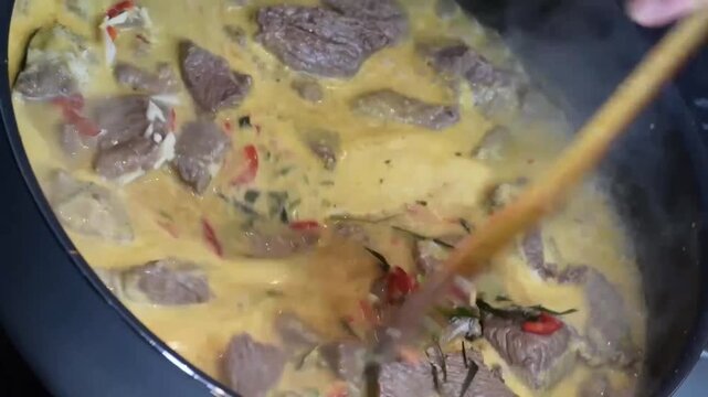 Beef curry simmering in a black pan as fresh red chilies, kaffir lime leaves, and garlic are stirred into the yellow sauce before covering with a glass lid