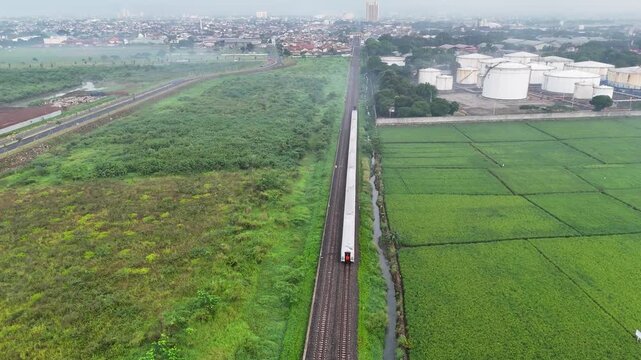 Aerial view of a train traveling on tracks through lush green rice fields near industrial storage tanks in Gedebage, Bandung, West Java, Indonesia.