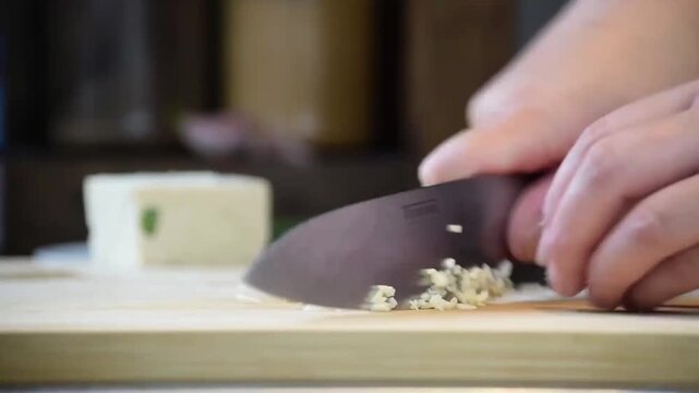 Hands mincing fresh garlic on a wooden cutting board with a chef's knife during food preparation