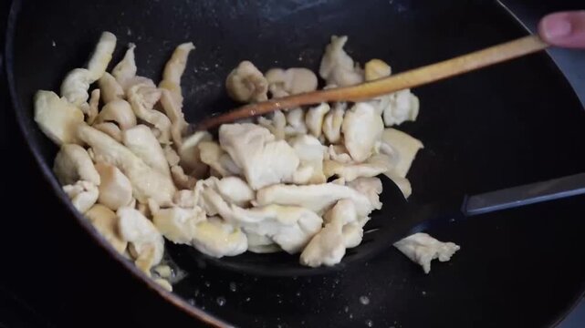 Chopped chicken pieces stir-frying in a black non-stick wok using wooden and slotted spatulas during home cooking