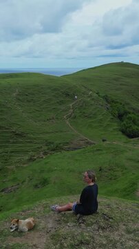 Woman standing by dog on cliff scanning wide coastal horizon, adjusting hair while wind sweeps over green hills, dog resting nearby, dramatic cloudscape and distant beaches frame reflective, vertical