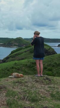 Woman standing by dog on cliff scanning wide coastal horizon, adjusting hair while wind sweeps over green hills, dog resting nearby, dramatic cloudscape and distant beaches frame reflective, vertical