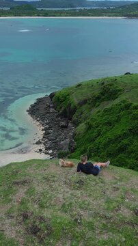 Woman lying beside dog on cliff above shallow turquoise lagoon, relaxed pose facing calm water, rocky inlet and distant shoreline visible, soft sky, casual summer attire, aerial camera,vertical video