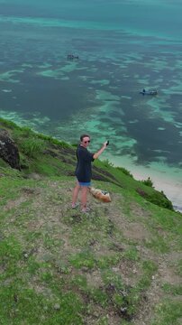 Caucasian woman photographing turquoise bay dog beside, smartphone raised for aerial frame, summer shorts and sandals, Bukit Merese, Lombok, Tanjung Ann, Indonesia, vertical video