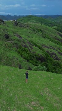 White woman walking hilltop overlooking bay solo photographer surveys sweeping green valleys and hidden sandy cove dramatic ridgelines aerial perspective adventurous tone, Bukit Merese, Lombok