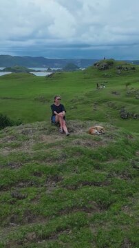 Woman lying beside dog on cliff above shallow turquoise lagoon, relaxed pose facing calm water, rocky inlet and distant shoreline visible, soft sky, casual summer attire, aerial camera,vertical video