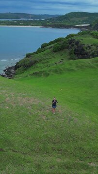 Woman gazing seaward. Solo woman pauses by ocean edge. Solitary woman stops at grassy cliff overlook. Individual female traveler gazes across calm ocean waters from rocky precipice, vertical video