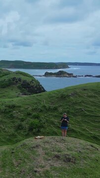 Caucasian woman cliff watching stormy bay dog resting beside, overcast sky and heavy clouds, green coastal slopes and rocky cove, smartphone held up to frame distant horizon, Lombok island