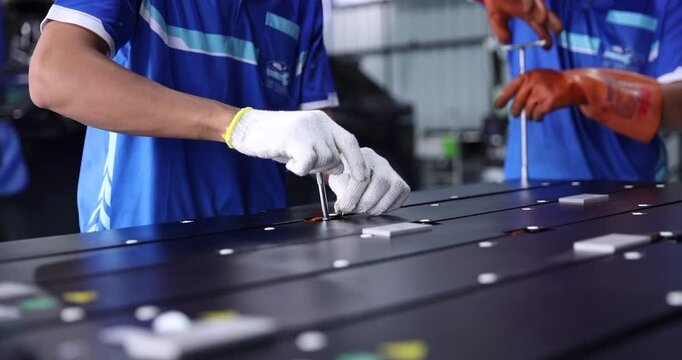 Two technicians in blue workwear use hand tools to tighten fasteners on electric vehicle battery modules. Close-up of gloved hands working together during battery pack assembly .
