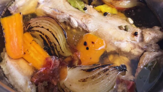Ingredients for a traditional hot soup base simmer in a pot as a chef cooks chicken stock with grilled onions, peppers, bay leaf, and fragrant spices into a healthy broth in a vertical video.