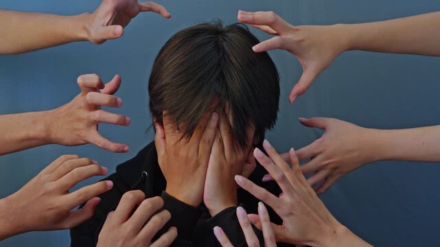 Distressed young asian man covering face while surrounded by multiple grabbing hands representing mental pressure 