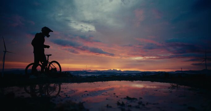 Silhouette of mountain bike rider cycling and standing off from bike, reflective water at colorful dusk sunset glow, centered symmetry
