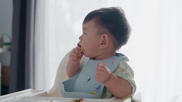 4K Asian baby boy eating boiled corn on high chair, BLW (baby-led weaning) concept, healthy solid food introducing to infant at home
