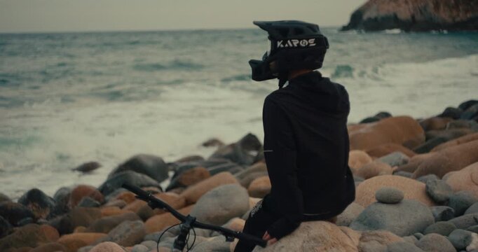Helmeted mountain bike rider standing among rocky boulders beside the ocean at dusk, cautious meditative moment in slow motion