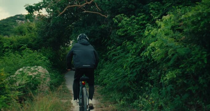 Mountain bike rider cycling away along narrow trail through dense green forest, rearview static on sandy path below trees