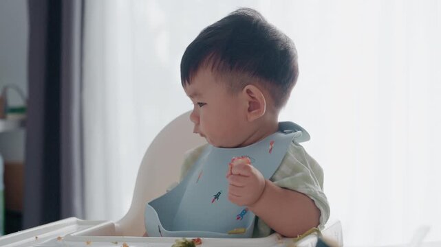 4K Asian baby boy eating boiled corn on high chair, BLW (baby-led weaning) concept, healthy solid food introducing to infant at home