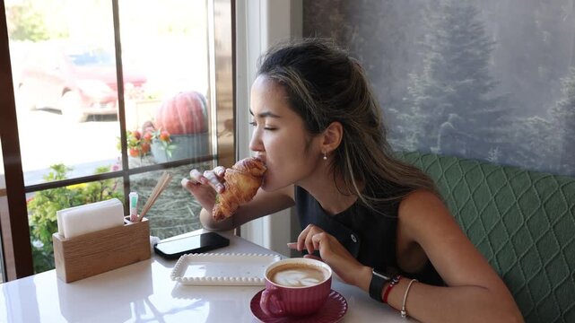 Asian woman eating croissant and enjoying coffee at a cozy cafe with bid window, casual breakfast in a modern restaurant interior, relaxed everyday dining lifestyle, bakery, hospitality