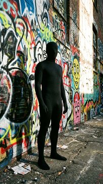 Anonymous figure in black spandex suit standing against colorful graffiti wall in urban alleyway environment