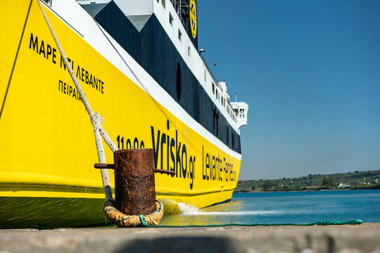 Low angle view of a rusty iron mooring bollard holding taut ropes from a yellow coastal passenger ferry docked alongside a stone quay in a Greek Mediterranean harbor.