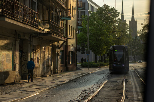 Poland, Wroclaw 2022 05 06: Street level shot of a modern tram moving on cobblestone tracks past old buildings and a walking man, illuminated by warm afternoon sunlight, horizontal view.