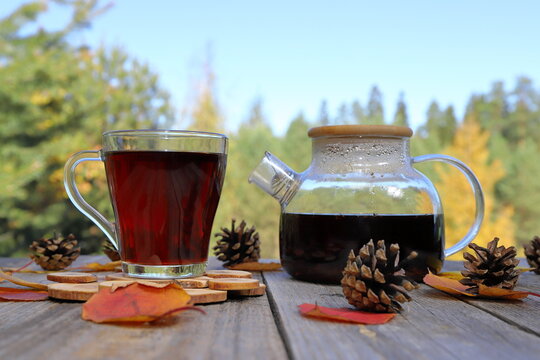 On a wooden table there are a glass mug of hot black tea and a glass teapot amidst foliage and pine cones, surrounded by an autumn forest landscape. 