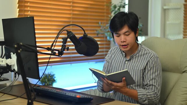 Young Asian man recording podcast at home studio, speaking into condenser microphone while holding notebook