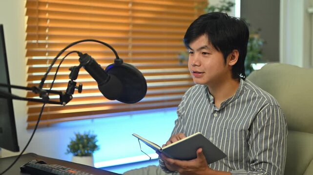 Young Asian man recording podcast at home studio, speaking into condenser microphone while holding notebook