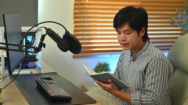 Young Asian man recording podcast at home studio, speaking into condenser microphone while holding notebook