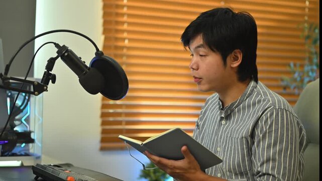 Young Asian man recording podcast at home studio, speaking into condenser microphone while holding notebook