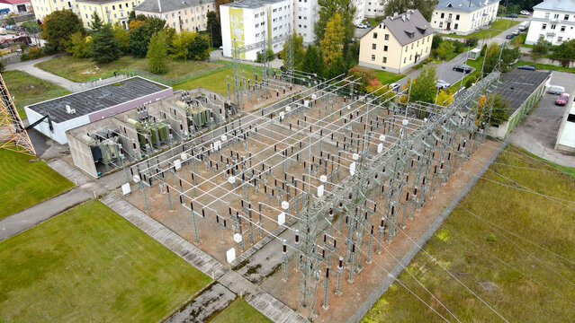 Aerial view showing an electrical substation with transformers, high voltage power lines, and multiple transmission towers distributing electricity to a surrounding urban landscape