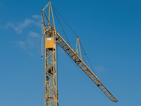Yellow tower crane cabin and luffing jib boom against clean blue sky. Heavy construction machinery, housing development and real estate industry concept.