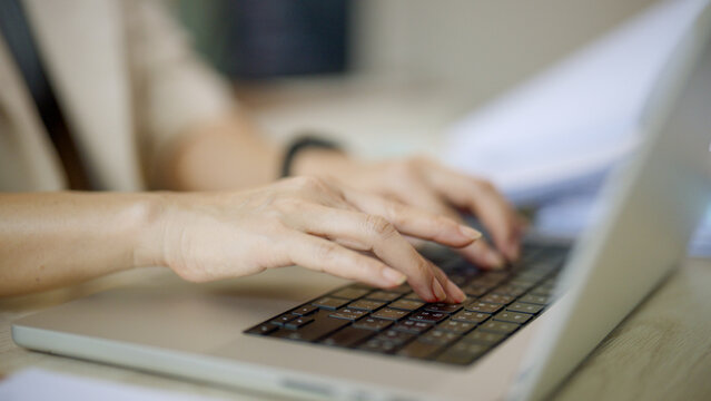 Soft focus shows hands typing on a laptop keyboard. Fingers move rhythmically over dark keys. Light wood desk holds the silver device. Focus draws attention to precision and activity