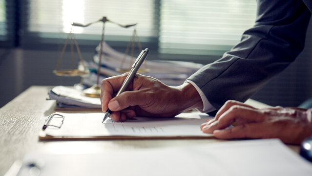 Hand writes legal document at desk. Scales of justice sit nearby. Focused, professional posture. Office light casts soft shadows. Ideal for legal or business contexts. Serious tone conveys formality