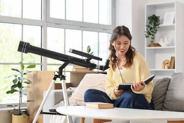 Fototapeta premium Astronomy hobby and education, young woman reading a book about space science with a telescope at home in a bright living room