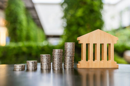 Banking institution and capital growth concept, classical wooden bank building model standing beside ascending coin stacks on table, symbolizing deposit savings, interest, and finance.